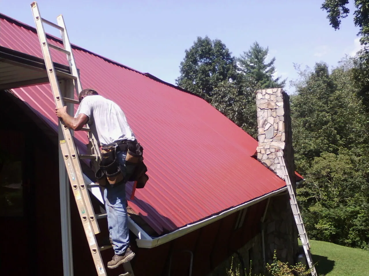 Roofer on a ladder beside a red metal roof and stone chimney — Soffit & Fascia Repair by Roofing Brothers in Quitman